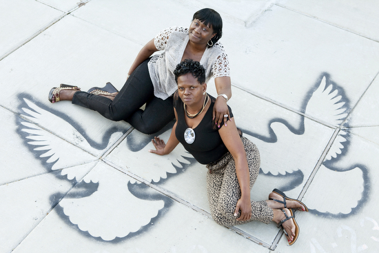 Martha Bullock (left) and Cathy Rickmon (right) sit on stencils of doves by artist Justin Kray on the corner of Baudin St. where their sons were killed on April 23, 2013. (Sabree Hill, UptownMessenger.com)